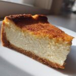A close-up slice of baked Protein Cheesecake with a thick, creamy white filling and a well-browned top, resting on a white plate.