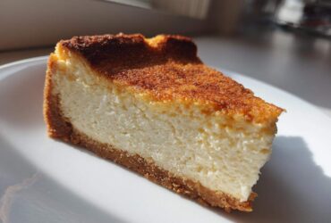A close-up slice of baked Protein Cheesecake with a thick, creamy white filling and a well-browned top, resting on a white plate.
