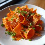 Close-up of a bright orange raw carrot salad made with shaved carrot ribbons, topped with sesame seeds and chopped green onions.