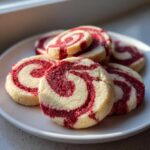 A stack of freshly baked red and white swirl pinwheel cookies resting on a white plate, beautifully lit by natural light.