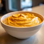 A close-up of a thick, vibrant orange beer cheese dip served in a white bowl, lightly dusted with paprika.