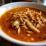 Close-up of a white bowl filled with rich, red broth and a generous topping of shredded chicken for chicken pozole.
