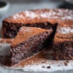 A close-up of a rich, dense slice of flourless chocolate torte, dusted heavily with cocoa powder.
