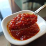 A spoonful of thick, vibrant red homemade pizza sauce being lifted from a small white bowl.