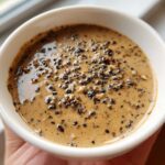 Close-up of a creamy, tan peppercorn sauce topped generously with cracked black peppercorns.