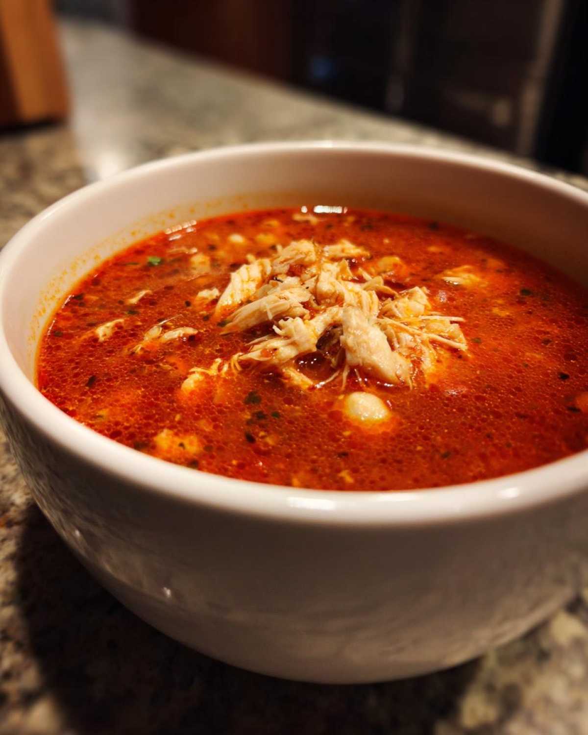 Close-up of a white bowl filled with vibrant red chicken pozole, topped with shredded chicken.