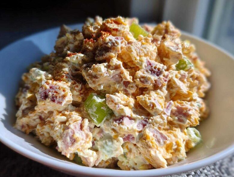 A close-up, bright photo of a creamy ham salad recipe served in a white bowl, topped with paprika.