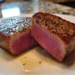 Two thick slices of perfectly seared tuna steak, showing a bright pink, medium-rare interior, seasoned with pepper.