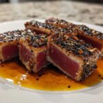 Close-up of perfectly seared sesame crusted tuna steak, sliced to show the rare red center, resting in sauce.