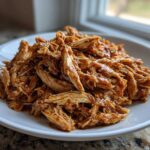 A mound of moist, shredded crockpot teriyaki chicken coated in a glossy sauce, served on a white plate.