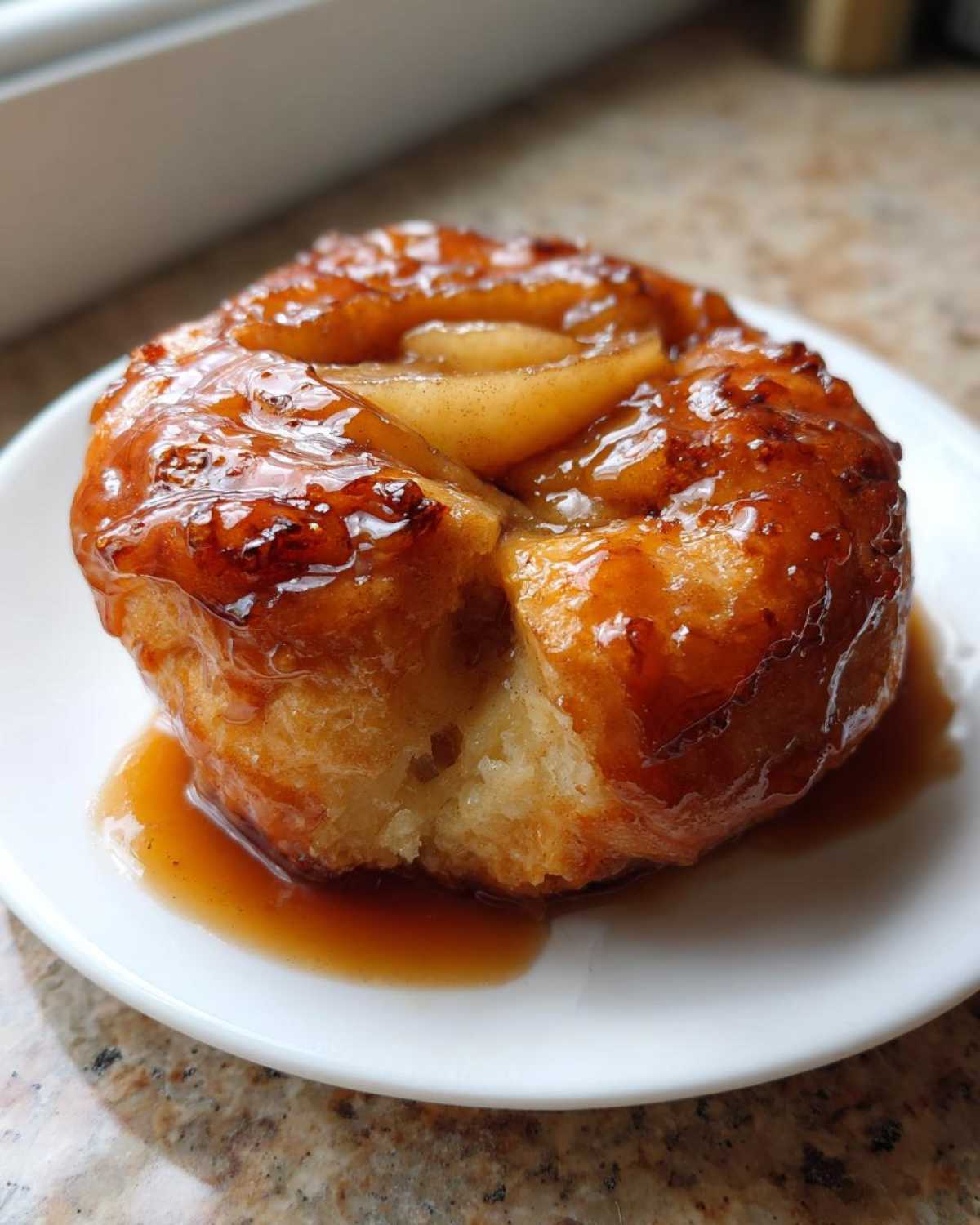 A close-up of a single, golden brown apple dumpling drenched in shiny caramel sauce, served on a white plate.