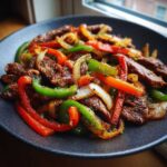 Close-up of sizzling steak fajitas featuring sliced beef, colorful bell peppers (red, green, orange), and onions served on a dark plate.