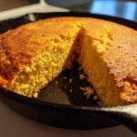 A golden brown, freshly baked skillet cornbread with one wedge cut out, showing the crumb texture.