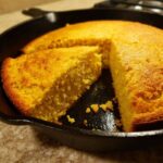 A close-up of golden, fluffy skillet cornbread baked in a cast iron pan with one wedge cut out.