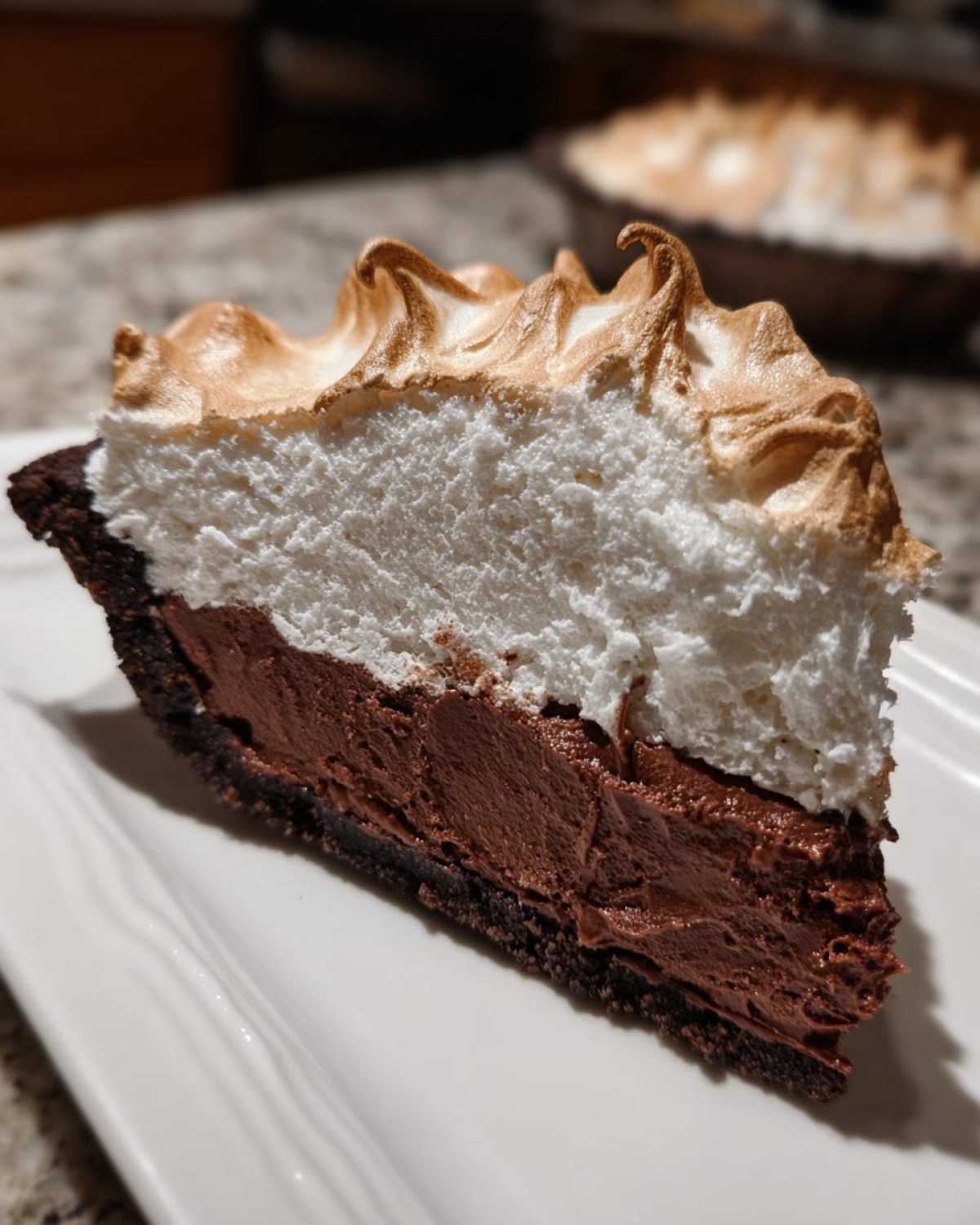 A thick slice of chocolate meringue pie showing a dark crust, rich chocolate filling, and tall, toasted meringue topping.