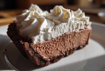 Close-up of a rich slice of chocolate mousse pie with a dark crust and generous topping of white whipped cream.