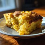 A golden slice of moist cornbread casserole studded with whole corn kernels, served on a white plate.