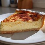 A close-up slice of creamy egg custard tart dusted with cocoa or cinnamon powder on a white plate.