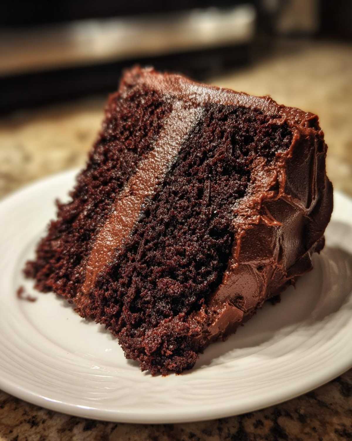 Close-up of a moist slice of homemade chocolate cake layered with thick chocolate frosting on a white plate.