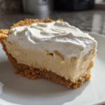 A thick slice of creamy peanut butter pie with a graham cracker crust and a generous topping of whipped cream.