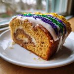 A close-up slice of homemade king cake featuring a cinnamon swirl, white icing, and purple, green, and gold Mardi Gras sprinkles.