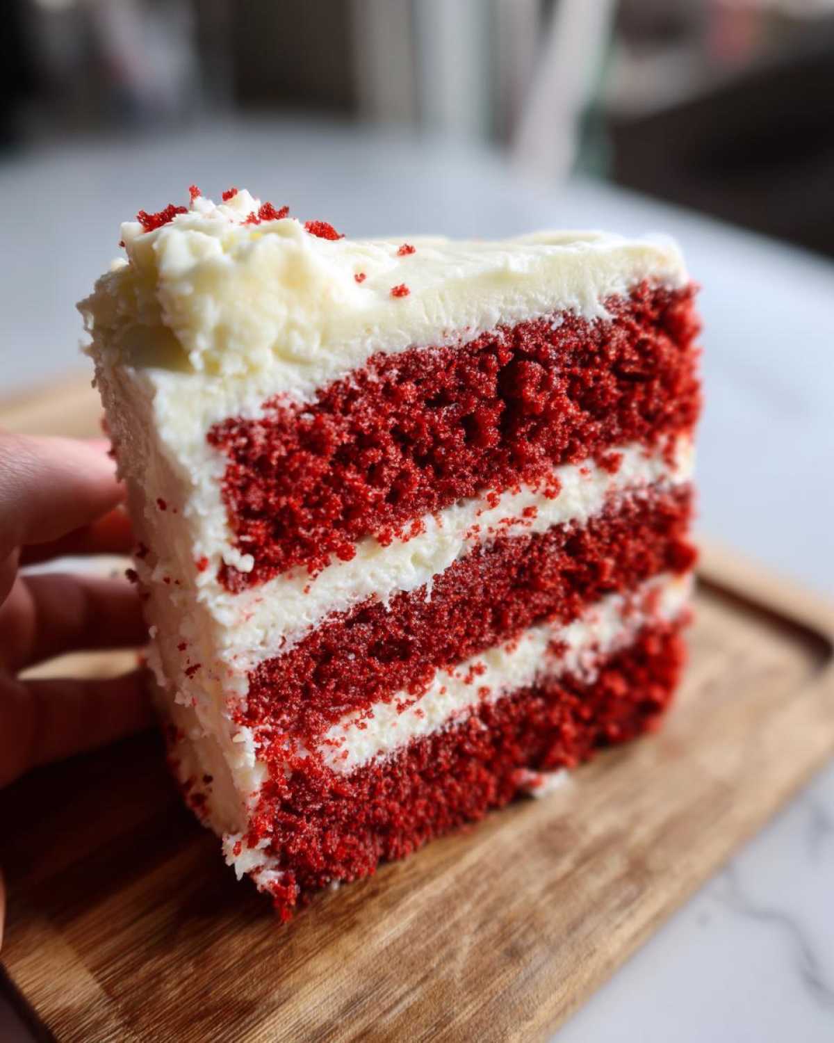 A tall slice of moist red velvet cake with three layers of deep red cake and white cream cheese frosting.