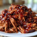 A close-up of tender, shredded Slow Cooker Pulled Pork generously coated in rich, glossy barbecue sauce on a white plate.