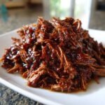 A close-up of tender, shredded Slow Cooker Pulled Pork coated in a rich, dark barbecue sauce on a white plate.