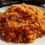 A close-up image showing a generous serving of vibrant, orange-red smoky jollof rice piled on a white plate.