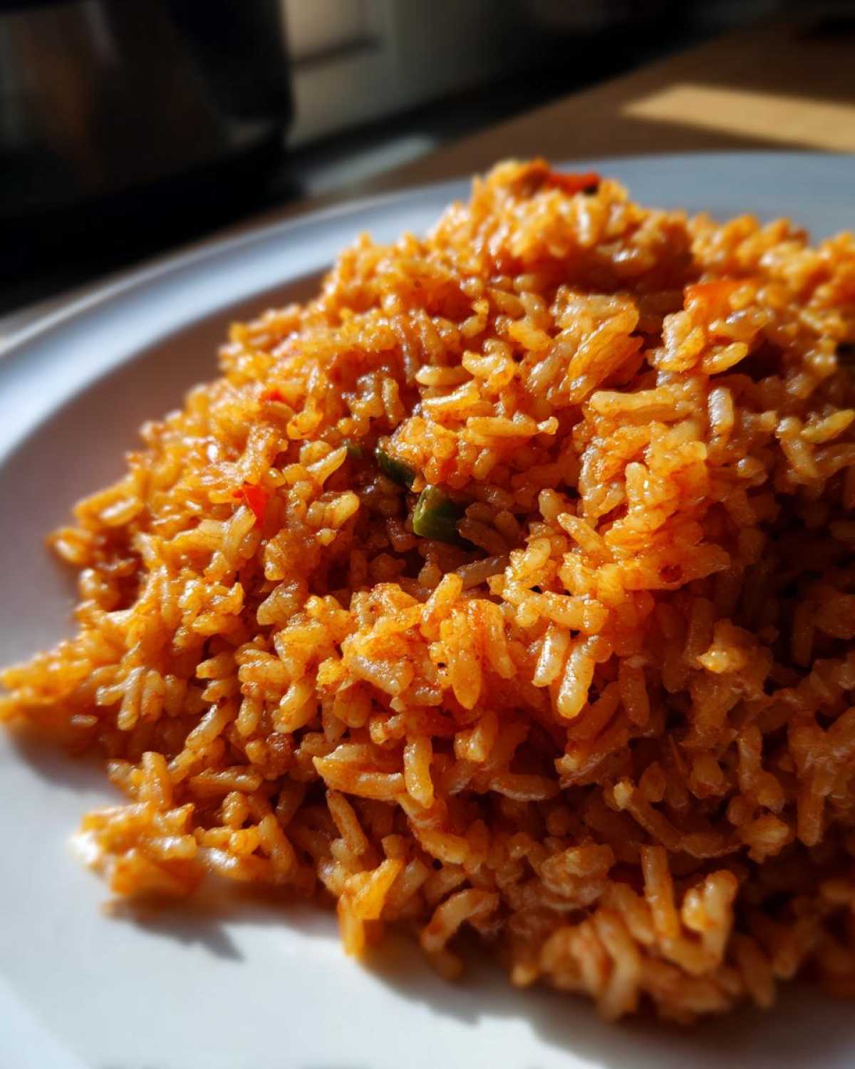 A close-up, sunlit shot of a mound of vibrant orange-red smoky jollof rice served on a white plate.