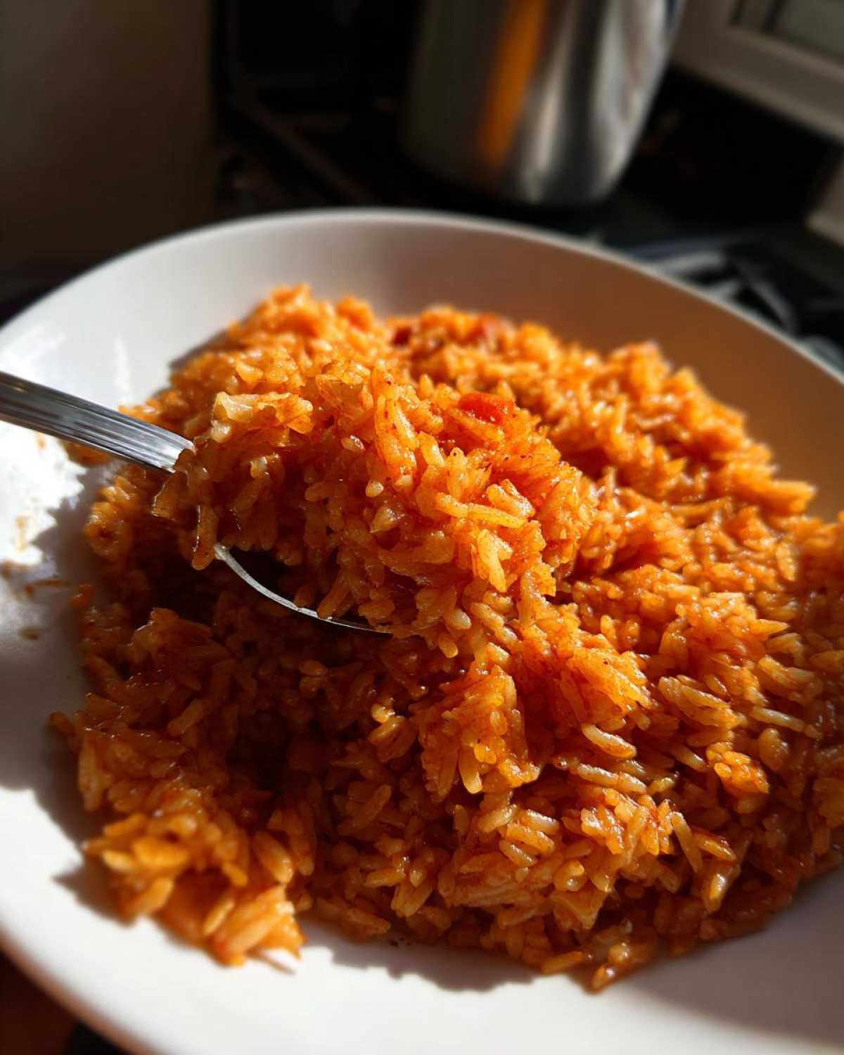 Close-up of a spoonful of vibrant, orange-red smoky jollof rice being lifted from a white bowl.