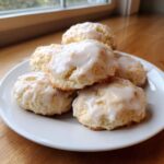 A close-up of several soft, round ricotta cookies piled on a white plate, generously drizzled with white glaze.