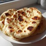 A stack of three freshly made, soft naan bread rounds with beautiful golden-brown charred spots, resting on a white plate.