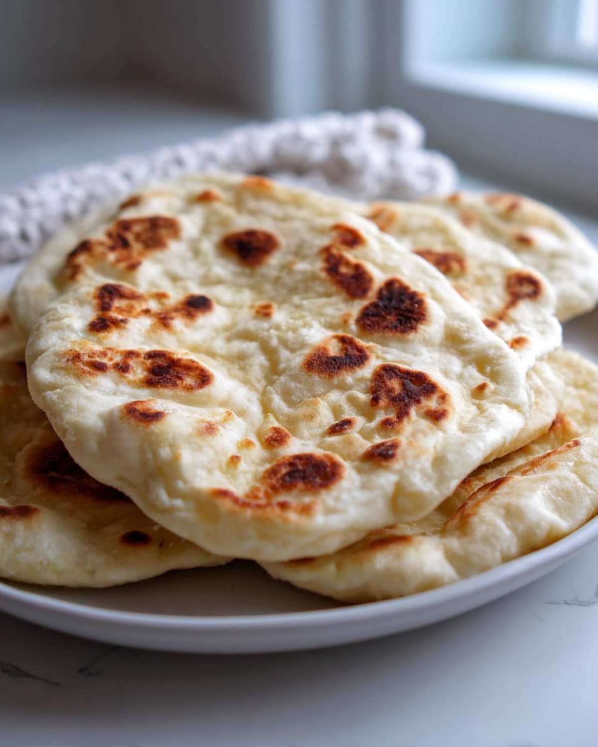 A stack of freshly made, soft naan bread with characteristic charred spots, resting on a white plate.