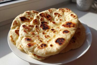 A stack of three freshly made, soft naan bread rounds with beautiful golden-brown charred spots, resting on a white plate.