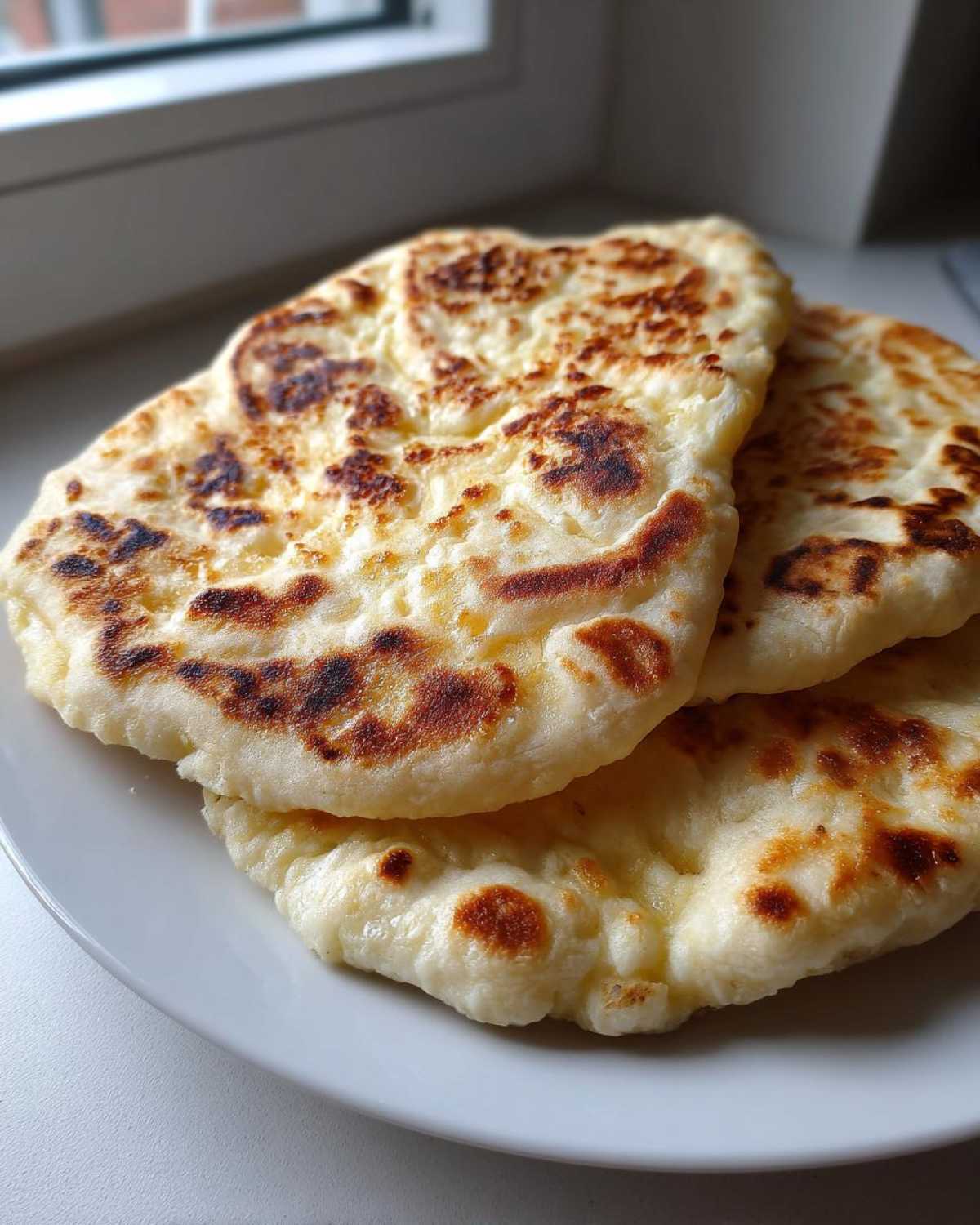 Three freshly cooked, soft naan bread recipe pieces stacked on a white plate with golden-brown charred spots.