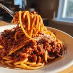 A fork lifting twirled spaghetti coated in thick bolognese sauce from a bowl, topped with grated cheese.
