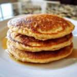 A close-up stack of three perfectly golden brown almond flour pancakes drizzled with syrup on a white plate.