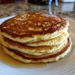 A delicious stack of four golden brown almond flour pancakes drizzled with syrup on a white plate.