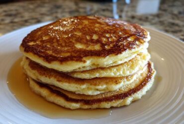 A delicious stack of four golden brown almond flour pancakes drizzled with syrup on a white plate.