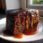 A close-up of a rich, dark slice of sticky toffee pudding being drenched in warm toffee sauce.