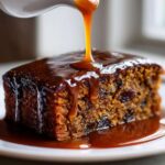 A close-up of a square slice of sticky toffee pudding recipe being drenched in rich caramel sauce from a white gravy boat.