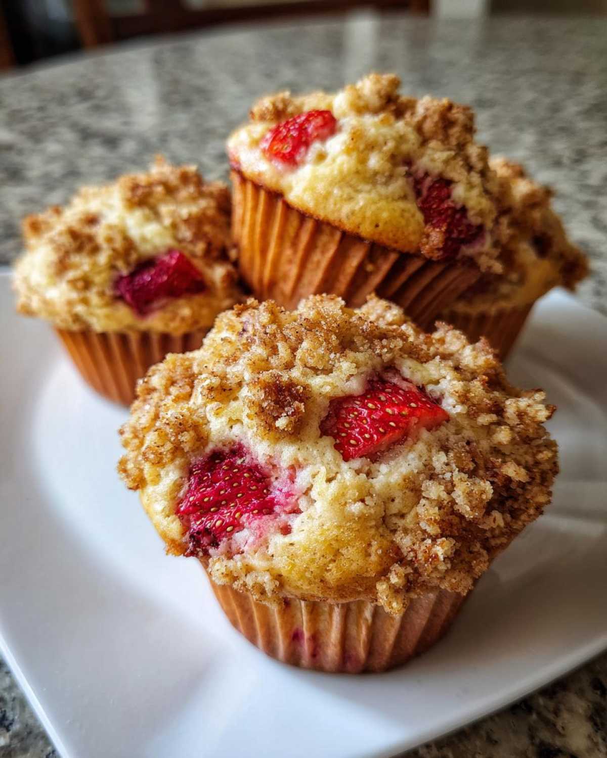 Three delicious strawberry muffins with a golden crumb topping arranged on a white plate.