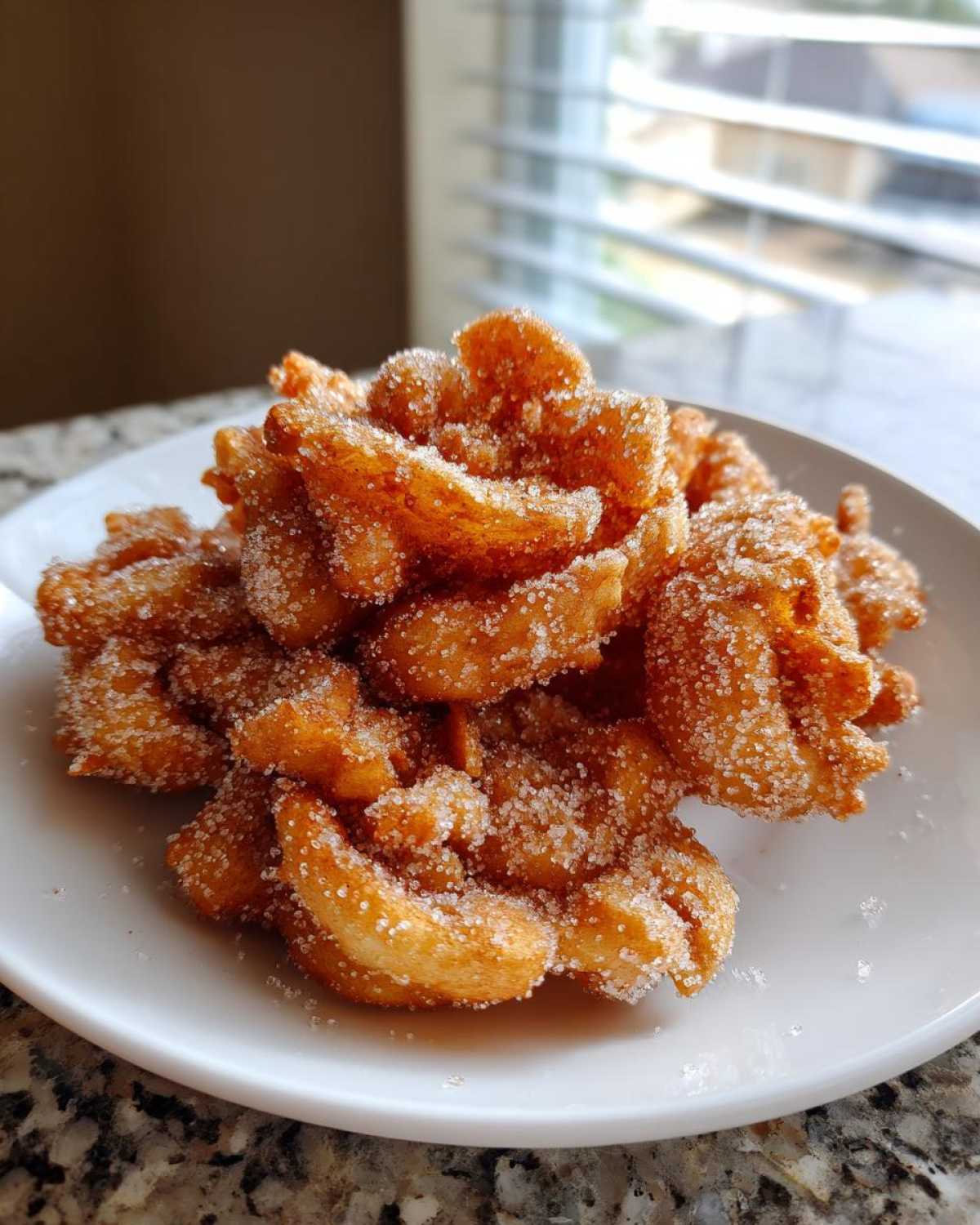 A close-up of a pile of golden, fried buñuelos heavily coated in granulated sugar, served on a white plate.