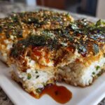Two squares of baked sushi bake casserole, topped with dark sauce, seaweed, and sesame seeds, served on a white plate.