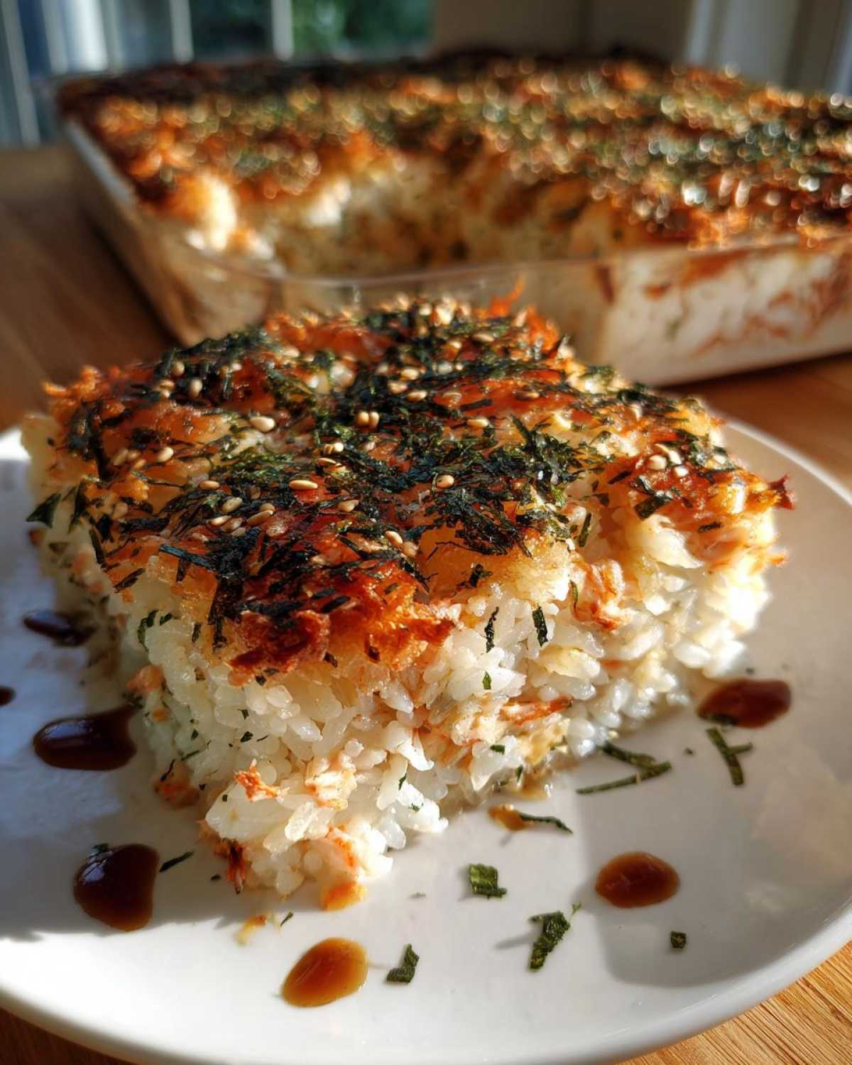A square slice of baked sushi bake casserole topped with furikake, served on a white plate with soy sauce drizzles.
