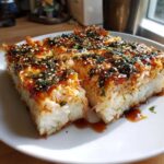 Three square slices of baked sushi bake casserole topped with crab, sauce, seaweed, and sesame seeds on a white plate.