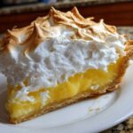 A tall slice of lemon meringue pie showing bright yellow filling and towering, toasted meringue peaks.