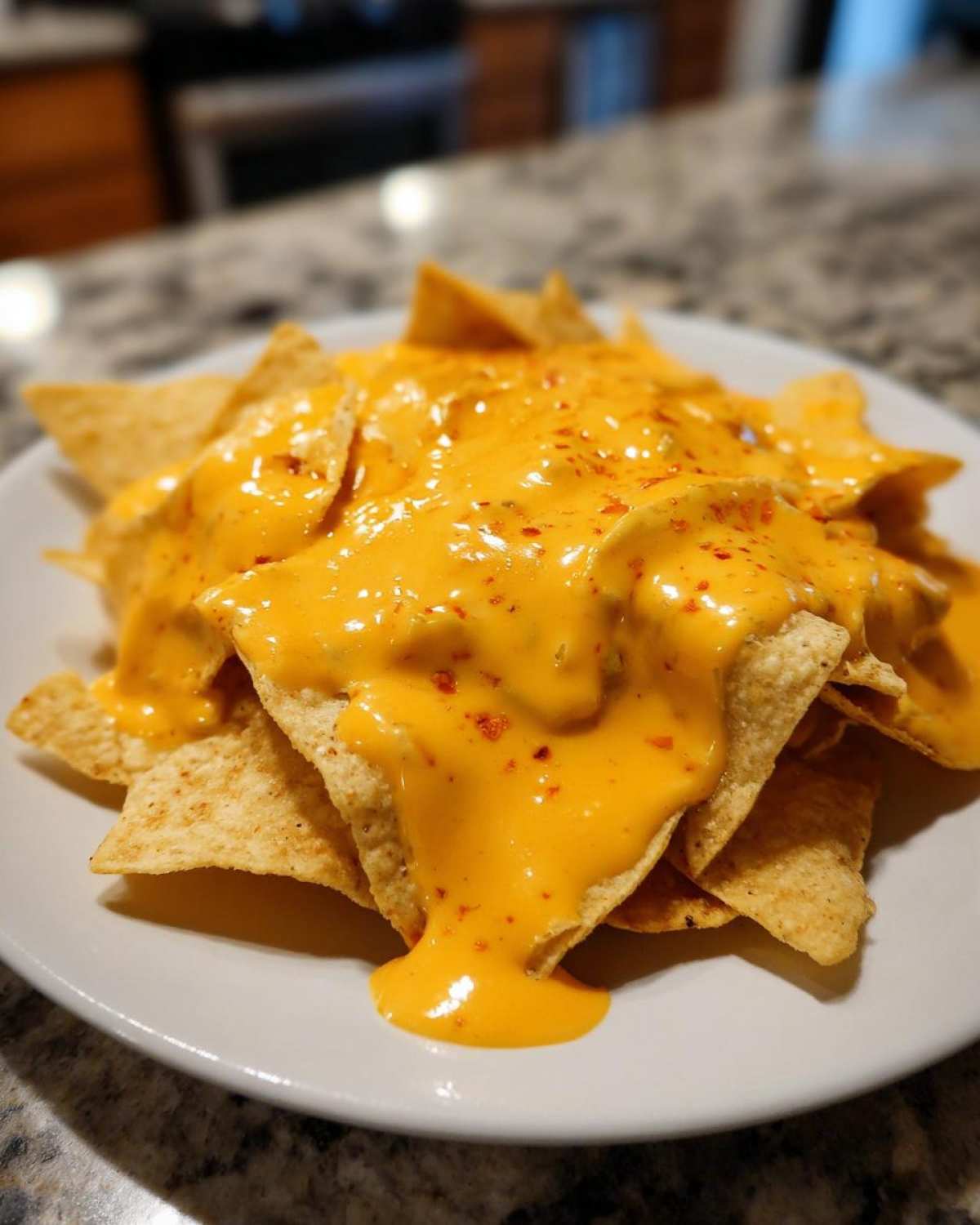 Close-up of tortilla chips generously covered in bright orange nacho cheese sauce, sprinkled with red pepper flakes.