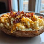 A close-up of a loaded twice-baked potato topped with melted cheddar cheese and crispy bacon bits.